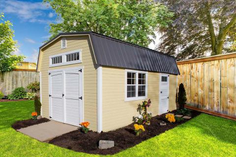 High barn shed with light yellow siding, white trim, white double doors, windows, a side door entrance, and a dark gray metal roof sitting in a fenced in backyard.